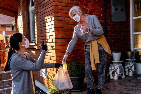 Woman with face mask delivering groceries to the front porch of elderly woman with face mask 
