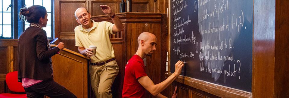 person writes equations on chalkboard while professor converses with student near podium
