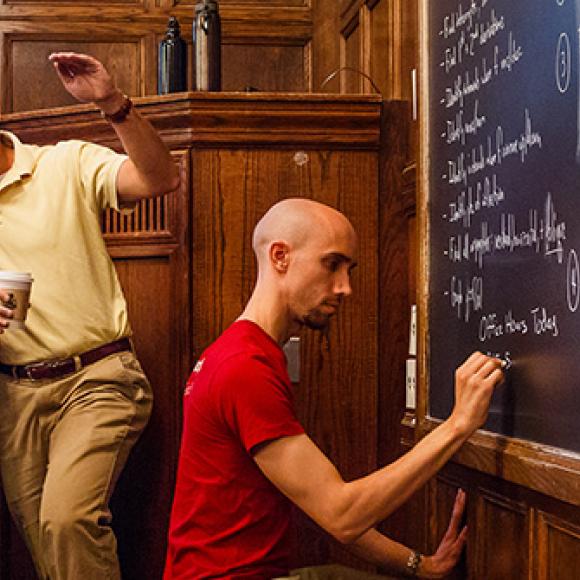 student writes on chalkboard while teacher and student talk at podium